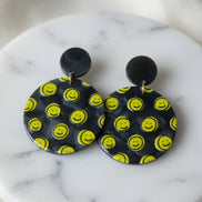 Avenue Zoe Happy Face Dangles earrings with cheerful face design and gold-tone hooks, hanging against a neutral background.