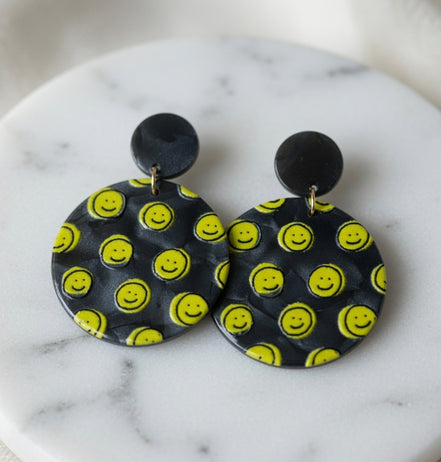Avenue Zoe Happy Face Dangles earrings with cheerful face design and gold-tone hooks, hanging against a neutral background.